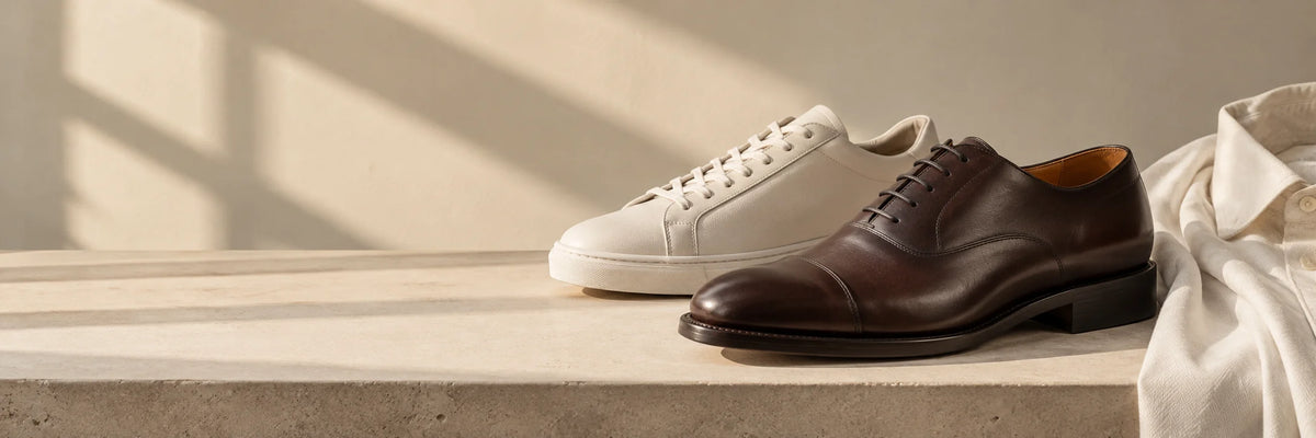 A brown cap-toe oxford and a white leather sneaker on a stone bench beside a cream shirt, in warm window light